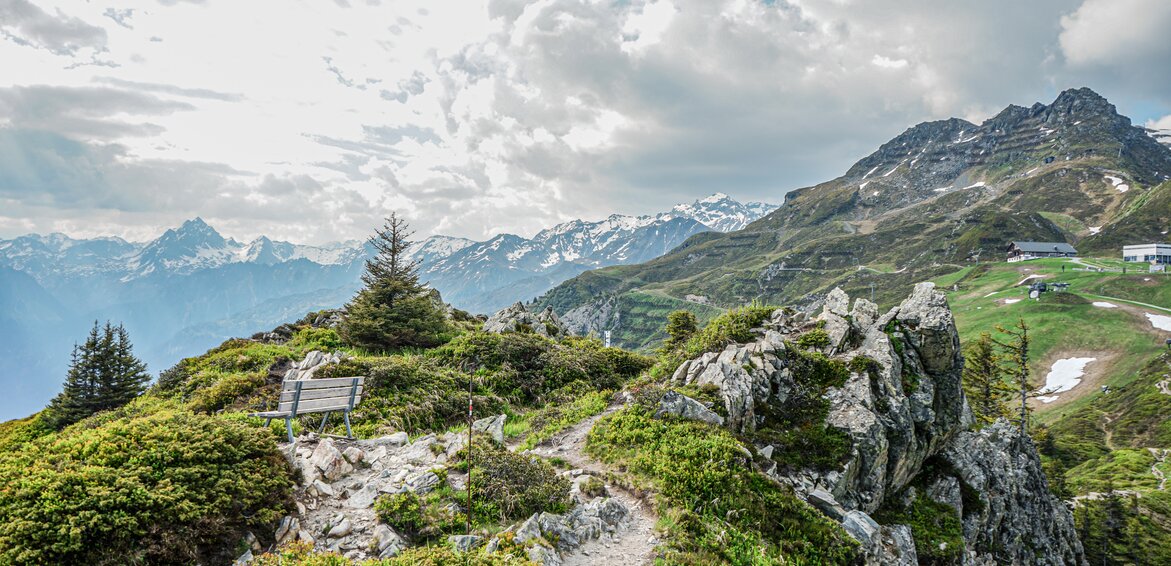 Ein Wanderweg an einem Grat mit Aussicht über die Berggipfel und einer Rastbank. | ©  Silvretta Montafon - Vanessa Strauch