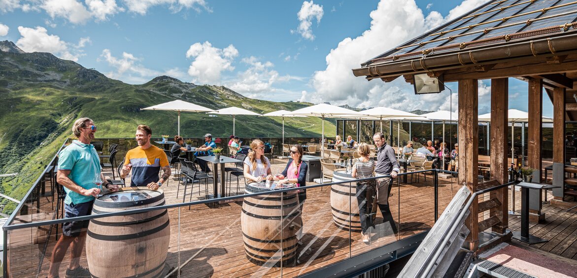Mehrere Personen stehen auf einer Terrasse mit Tischen und Weinfässern direkt in den Bergen der Silvretta Montafon. | © Silvretta Montafon - Stefan Kothner
