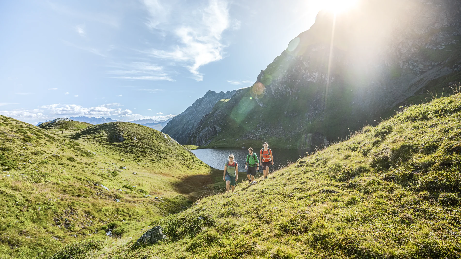 Drei Personen wandern bei Sonnenschein im Sommer in der Nähe eines Bergsees umringt von hohen Bergen. | © Silvretta Montafon - Stefan Kothner
