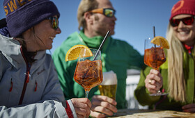 Zwei Damen und ein Herr stoßen auf der Bella Nova Terrasse mit Bier und Aperol auf den tollen Tag an