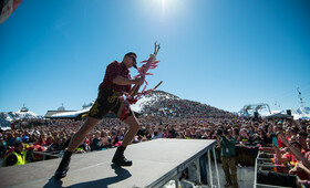 Andreas Gabalier steht mit Mikrofon in der Hand auf der Bühne vor seinen Fans unter blauen Himmel und Sonnenschein
