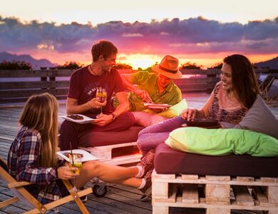 Vier junge Erwachsene sitzen gemütlich mit Essen und Trinken in der Hand auf der Hochjoch Terrasse