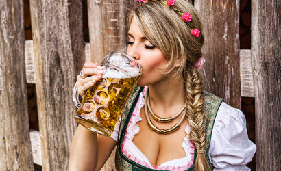 Eine junge Dame mit Blumenkranz im Haar lehnt an einer Holzwand und trinkt aus ihrem Maß Bier