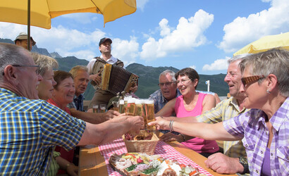 Eine Seniorengruppe sitzt auf der Terrasse, stoßt mit ihren Biergläsern an und isst Brettljause