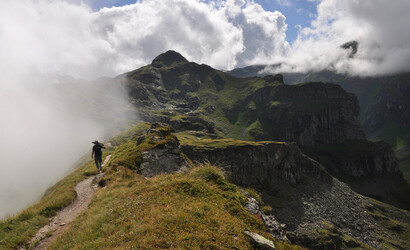 Ein Mann mit einem Kunstwerk auf den Schultern läuft einen Wanderweg in den Bergen entlang