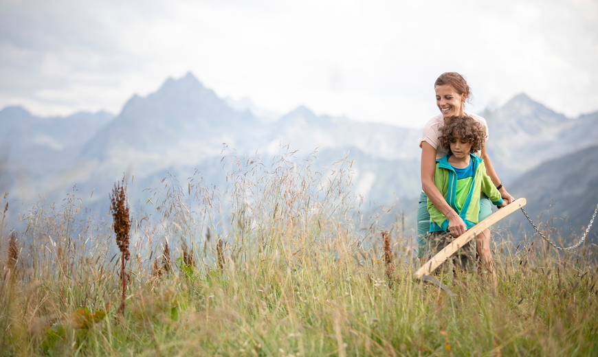 Eine Mama und ihr Sohn mähen das hohe Gras bei der Spielstation Alpenwirtschaft in der Alpenwelt Nova