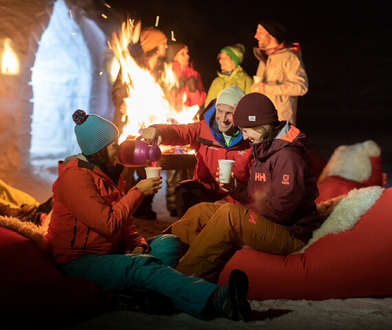 Zwei Personen sitzen in roten Sitzsäcken und trinken warme Getränke vor einem Feuer im Igludorf