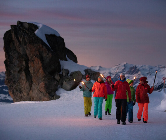 Eine Menschengruppe in Skibekleidung wandert mit Fackeln durch den Schnee in der Abenddämmerung in der Silvretta Montafon 