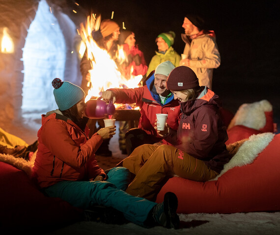 Zwei Personen sitzen in roten Sitzsäcken und trinken warme Getränke vor einem Feuer im Igludorf