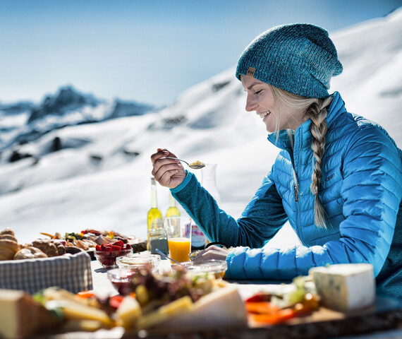Eine Frau sitzt an einem Tisch in den Bergen im Winter und vor ihr ist ein Frühstücksbuffet aufgebaut