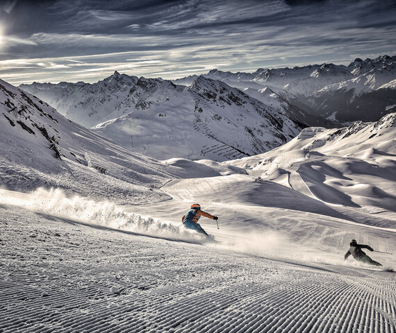 Zwei Skifahrer sausen die frisch präparierten Pisten hinunter mit Bergpanorama und Sonnenschein im Hintergrund