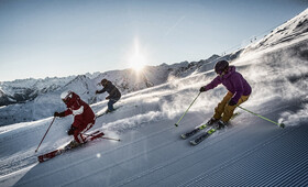 Ein Skiguide und zwei Personen in Skikleidung und Helm sausen die frische Piste im Sonnenschein hinunter in den Bergen