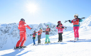 Eine Gruppe von Kinder steht in der Reihe nebeneinander auf der Piste und beobachtet den Skilehrer im Sonnenschein