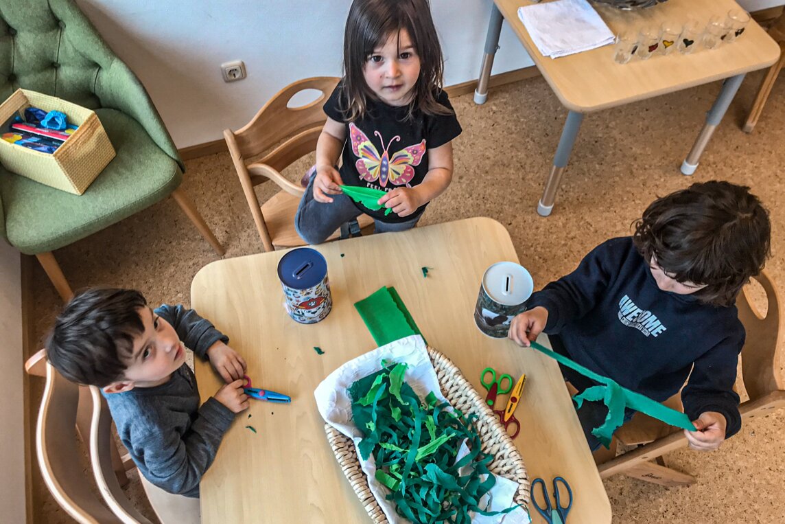 Drei Kinder sitzen an einem Tisch in einer Kinderbetreuung und Basteln etwas mit grünen Papierstreifen. | © Silvretta Montafon - Katharina Liebe