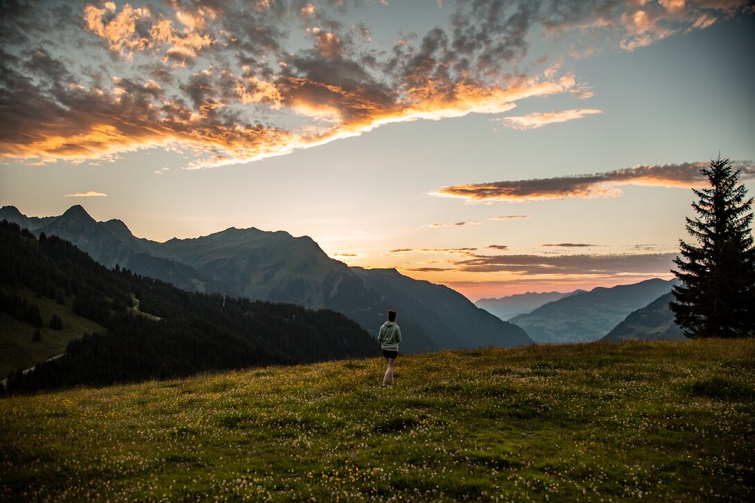 Eine Person steht in der Wiese am Berg und genießt das Abendlicht im Bergdorf in der SIlvretta Montafon. | ©  Silvretta Montafon - Friederike Weber
