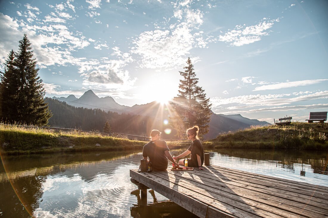 Abendfahrt Garfrescha - Silvretta Montafon | © Silvretta Montafon - Friederike Weber Zwei Personen picknicken auf einem Steg in der Abendsonne am See im Alpendorf Garfrescha. | © Silvretta Montafon - Friederike Weber