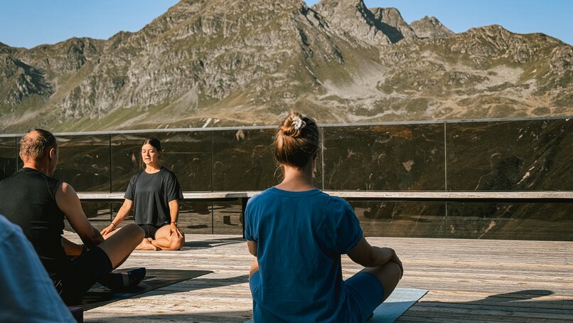 Eine Gruppe von Personen sitzt auf einer Terrasse bei Morgensonne und macht Yoga. | © Silvretta Montafon - Vanessa Strauch