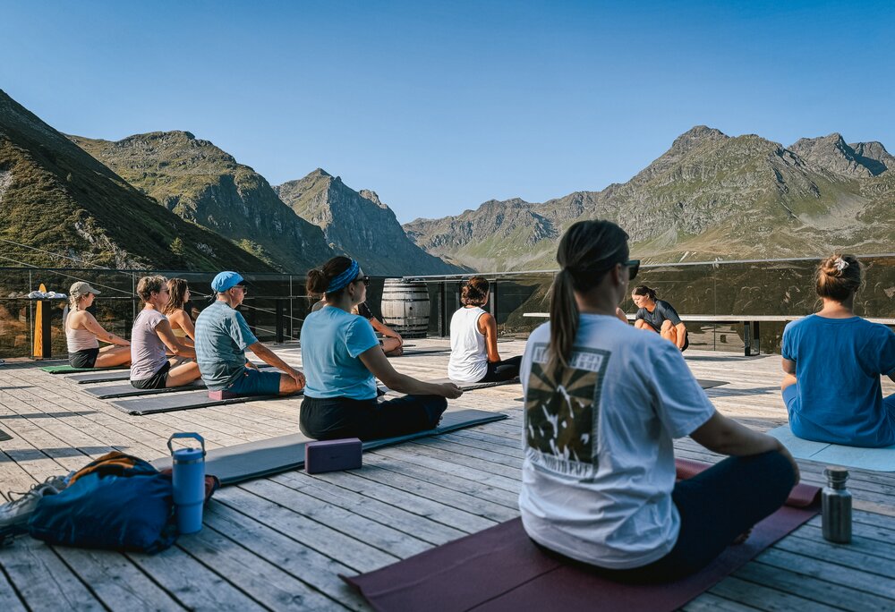 Eine Gruppe von Personen sitzt auf einer Terrasse bei Morgensonne und macht Yoga. | © Silvretta Montafon - Vanessa Strauch