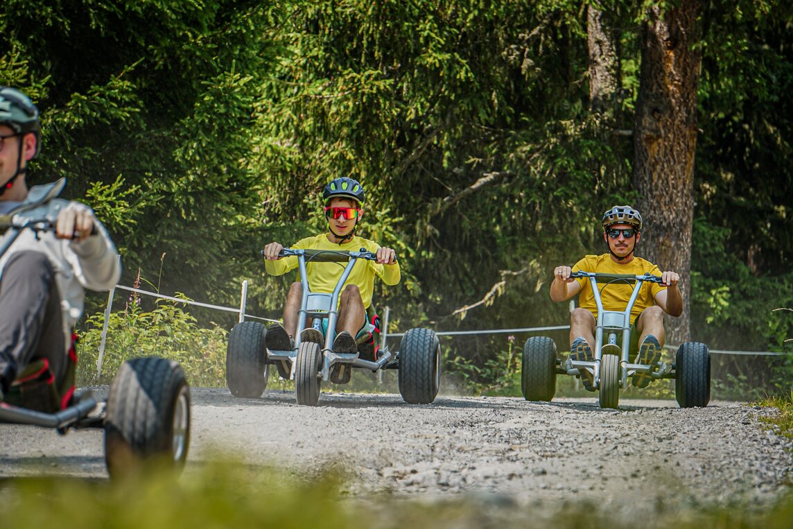 Drei junge Männer fahren auf Mountaincarts einen Güterweg im Wald entlang. | © Silvretta Montafon - Vanessa Strauch