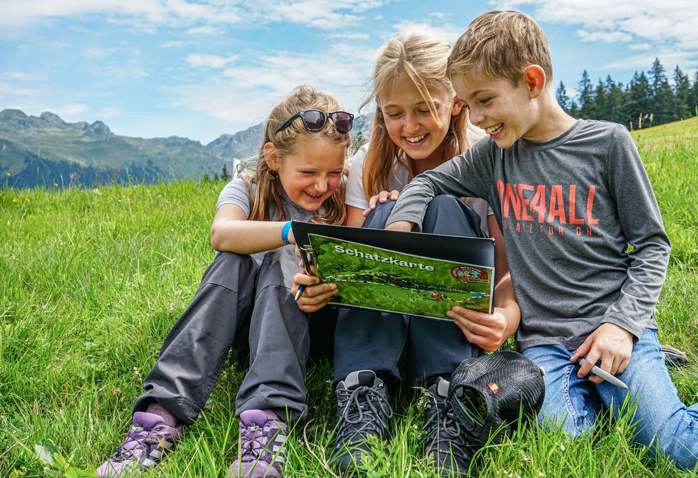 Drei Kinder sitzen auf einer Wiese und lesen eine Schatzkarte in der Silvretta Montafon. | © Silvretta Montafon - Vanessa Strauch