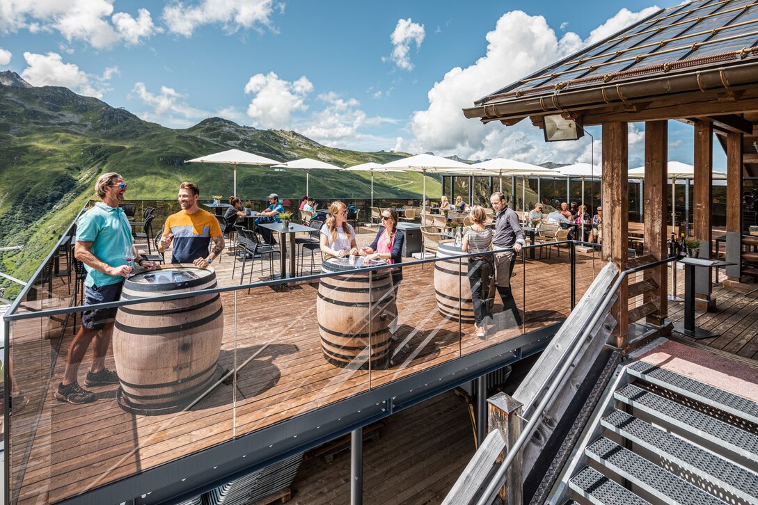 Mehrere Personen stehen auf einer Terrasse mit Tischen und Weinfässern direkt in den Bergen der Silvretta Montafon. | © Silvretta Montafon - Stefan Kothner