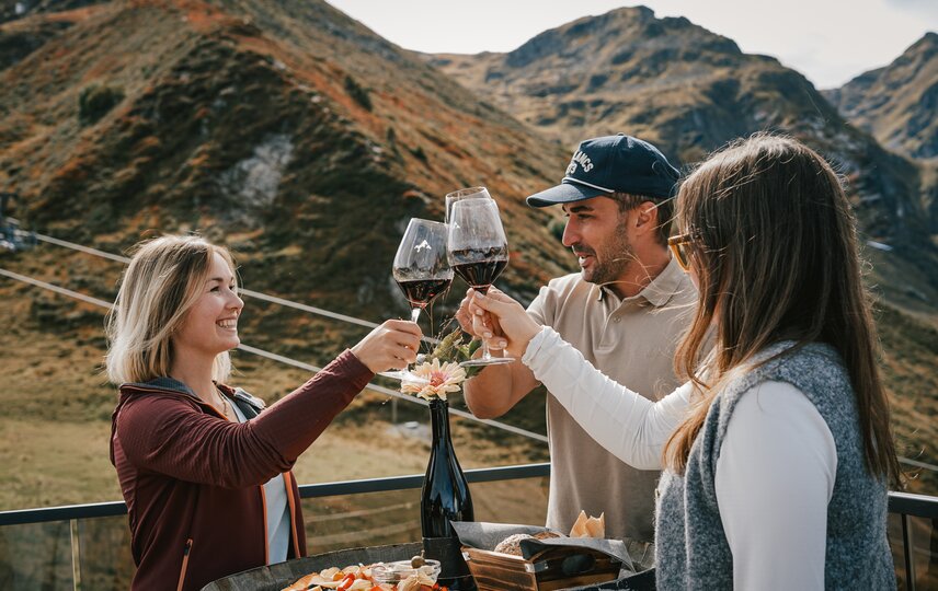 Drei Personen stehen an einem Weinfass mit Käse und Brot und stoßen mit einem Glas Rotwein an. | © Silvretta Montafon - Vanessa Strauch