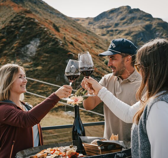 Drei Personen stehen an einem Weinfass mit Käse und Brot und stoßen mit einem Glas Rotwein an. | © Silvretta Montafon - Vanessa Strauch