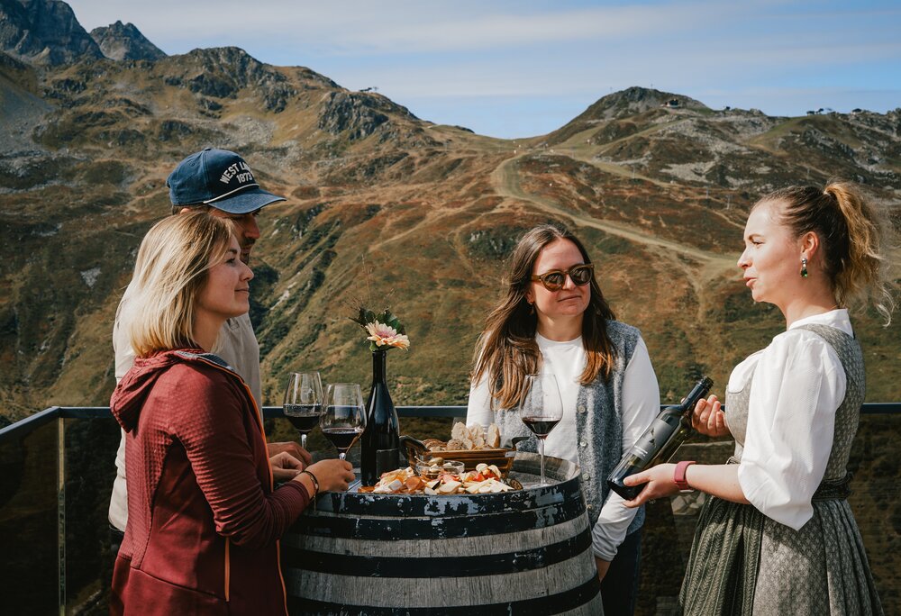 Drei Personen stehen an einem Weinfass mit einem Glas Rotwein an und eine Kellerin im Dirndl erklärt den Wein. | © Silvretta Montafon - Vanessa Strauch