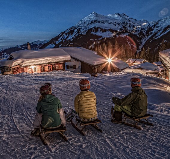 Drei Personen sitzen auf einem Schlitten in einem Bergdorf am Abend in der Silvretta Montafon. | © Silvretta Montafon - Daniel Zangerl