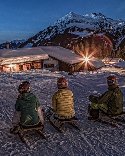 Drei Personen sitzen auf einem Schlitten in einem Bergdorf am Abend in der Silvretta Montafon. | © Silvretta Montafon - Daniel Zangerl