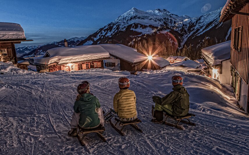 Drei Personen sitzen auf einem Schlitten in einem Bergdorf am Abend in der Silvretta Montafon. | © Silvretta Montafon - Daniel Zangerl
