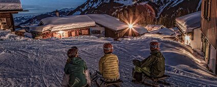 Drei Personen sitzen auf einem Schlitten in einem Bergdorf am Abend in der Silvretta Montafon. | © Silvretta Montafon - Daniel Zangerl