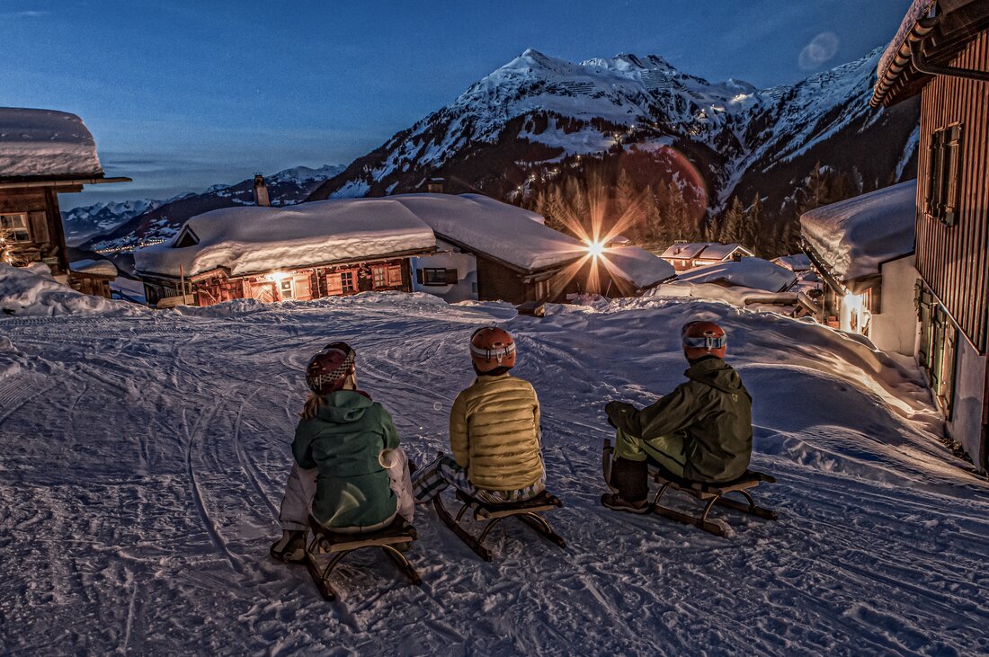 Drei Personen sitzen auf einem Schlitten in einem Bergdorf am Abend in der Silvretta Montafon. | © Silvretta Montafon - Daniel Zangerl