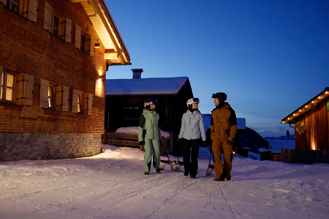 Drei Personen ziehen einen Rodel in einem verschneiten Bergdorf am Abend. | © Silvretta Montafon - Niklas Kirchler