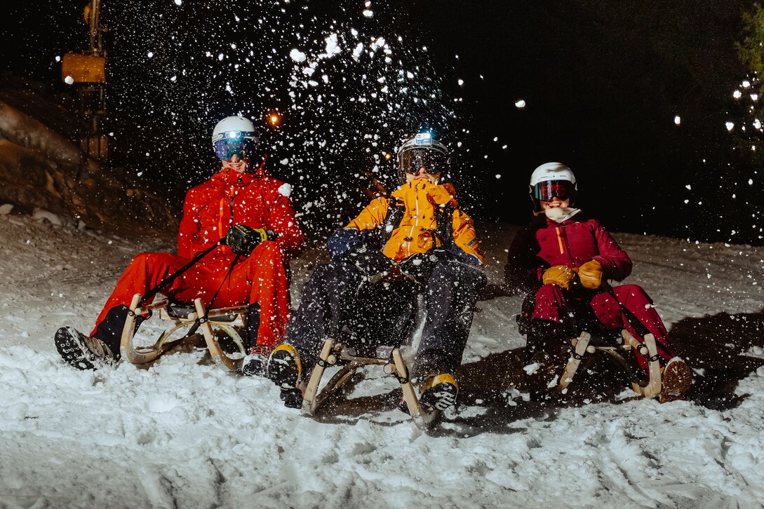 Drei Personen rodeln auf einem Schlitten und der Schnee spritzt in die Kamera. | © Silvretta Montafon - Marie Schilcher