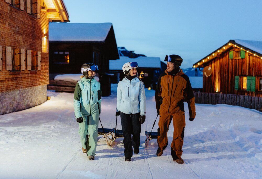 Drei Personen in Skikleidung ziehen einen Rodel durch ein verschneites Bergdorf am Abend. | © Silvretta Montafon - Marie Schilcher