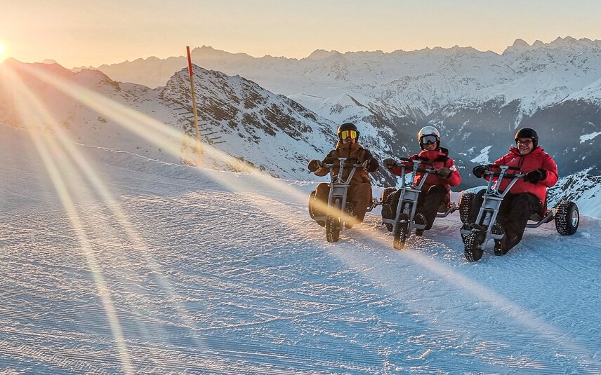 Drei Männer sitzen auf einem Mountain Cart im Schnee bei Sonnenaufgang auf der Skipiste. | © Silvretta Montafon