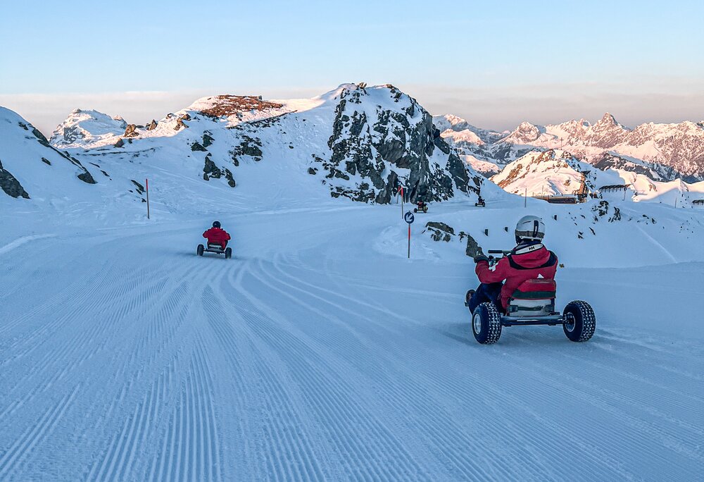Zwei Personen fahren auf einem Mountain Cart im Schnee bei Sonnenaufgang auf der Skipiste. | © Silvretta Montafon