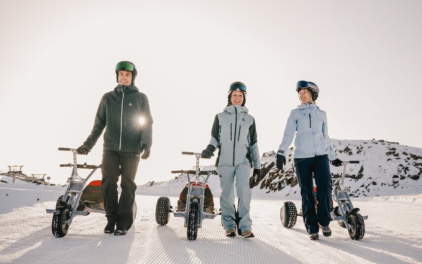 Drei Personen laufen mit einem Mountaincart auf einer Skipiste am Morgen bei Sonnenaufgang. | © Silvretta Montafon - Niklas Kirchler