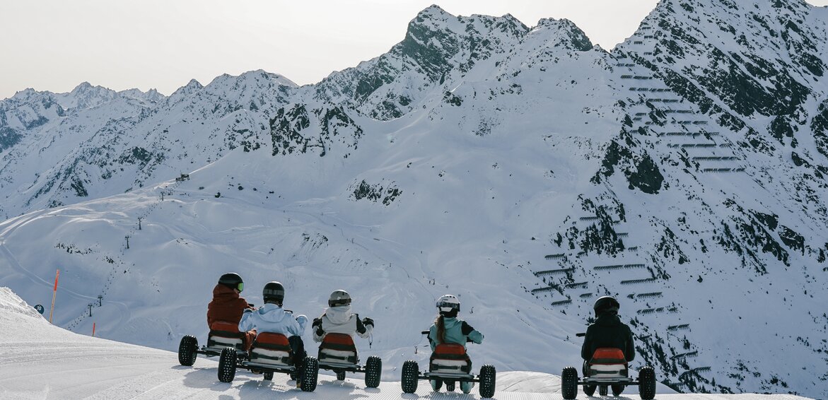 Fünf Personen auf einem Mountaincart auf einer Skipiste am Morgen bei Sonnenaufgang und Blick auf die verschneiten Berge. | © Silvretta Montafon - Niklas Kirchler