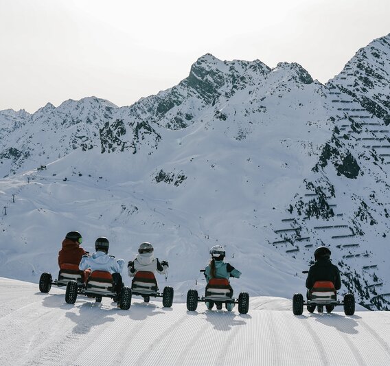 Fünf Personen auf einem Mountaincart auf einer Skipiste am Morgen bei Sonnenaufgang und Blick auf die verschneiten Berge. | © Silvretta Montafon - Niklas Kirchler