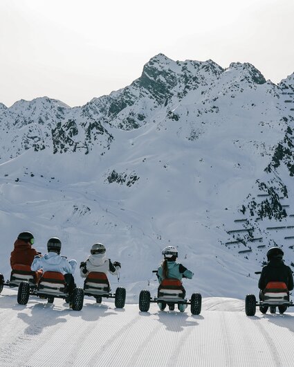 Fünf Personen auf einem Mountaincart auf einer Skipiste am Morgen bei Sonnenaufgang und Blick auf die verschneiten Berge. | © Silvretta Montafon - Niklas Kirchler