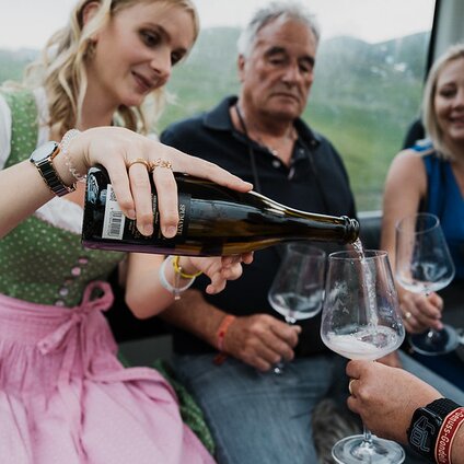 Eine junge Frau in Tracht schenkt ein Glas Wein in einer Gondel für eine Dame ein | © Silvretta Montafon - GOODGUYPAEZ