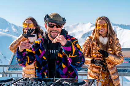 Sebastian Bronk am DJ Pult mit zwei Tänzerinnen in Orange im Hintergrund | © Silvretta Montafon - Sebastian Bronk