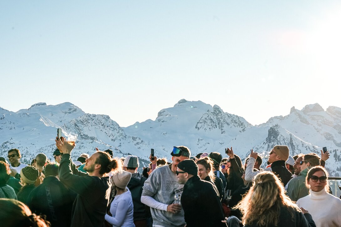 Viele feiernde Gäste auf einer Terrasse bei Sonnenuntergang und Panorama auf die verschneiten Gipfel. | © Silvretta Montafon - Vanessa Strauch