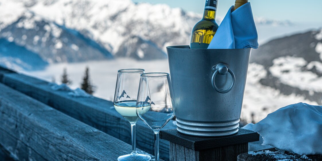 Zwei Gläser Wein und ein Kühler mit zwei flaschen Weißwein stehen auf der Terrasse des Kapellrestaurants. | © Silvretta Montafon - Vanessa Strauch