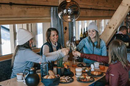Vier Frauen sitzen an einem reichlich gedeckten Frühstückstisch und stoßen mit einem Glas Sekt an. | © Silvretta Montafon - Vanessa Strauch