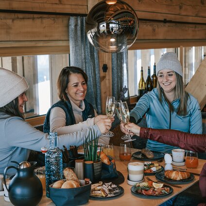Vier Frauen sitzen an einem reichlich gedeckten Frühstückstisch und stoßen mit einem Glas Sekt an. | © Silvretta Montafon - Vanessa Strauch