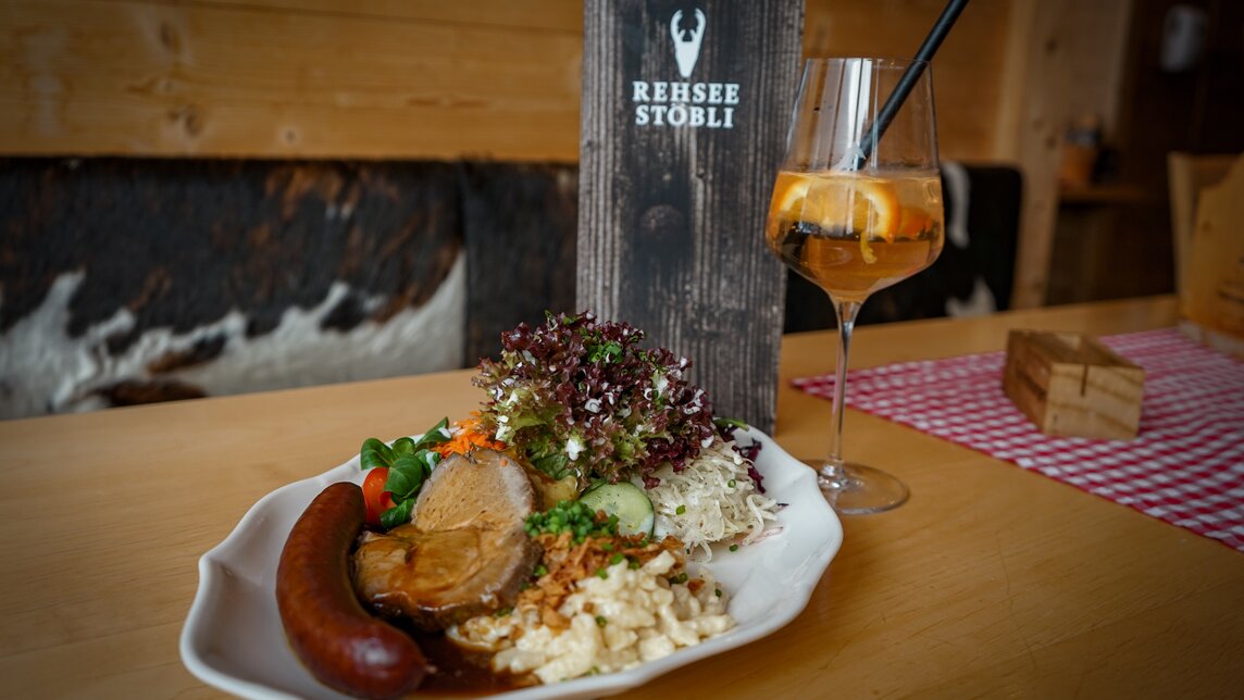 Ein Teller mit Hauswurst, Fleisch, Käsespätzli und Salat, daneben ein Aperol Spritz. | © Silvretta Montafon - Vanessa Strauch