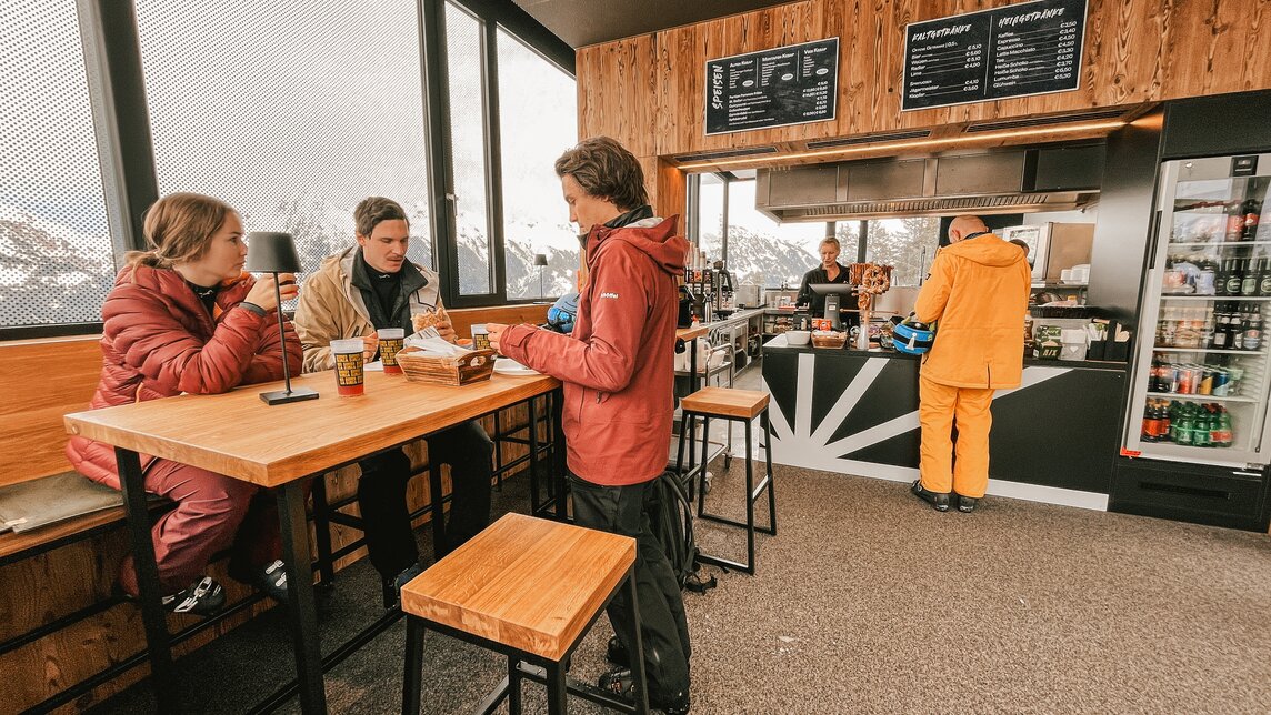 Drei Personen in der Sonnenbarga an einem Stehtisch und eine Person bestellt etwas an der Theke. | © Silvretta Montafon - Vanessa Strauch